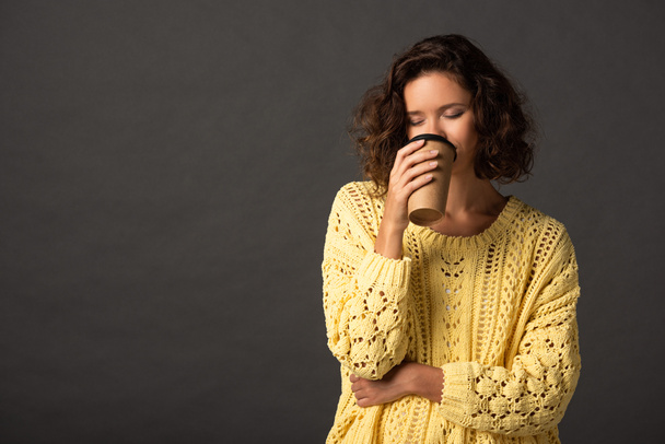 curly woman in yellow knitted sweater with closed eyes holding coffee to go on black background - Foto, immagini