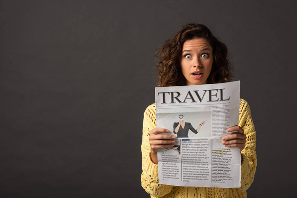 shocked curly woman in yellow knitted sweater holding travel newspaper on black background - Photo, Image