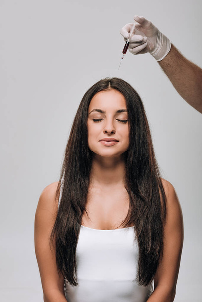Kosmetikerin in Latex-Handschuhen macht Kopfhaut Mesotherapie für brünette schöne Frau mit langen Haaren isoliert auf grau - Foto, Bild