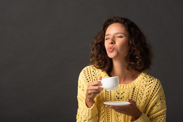 curly woman in yellow knitted sweater pouting lips while holding cup of coffee and saucer on black background - Photo, Image