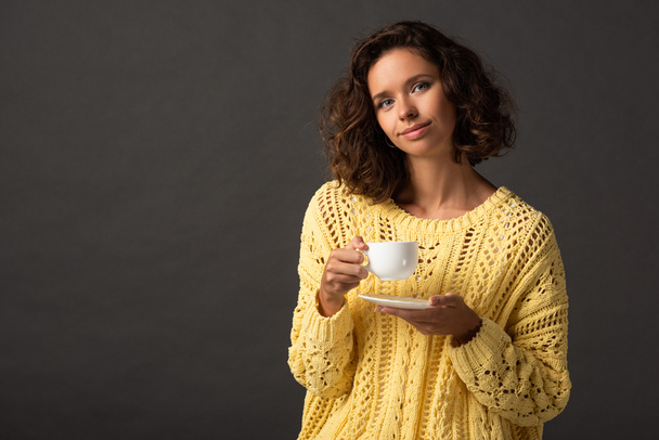 smiling curly woman in yellow knitted sweater holding cup of coffee and saucer on black background - Foto, imagen