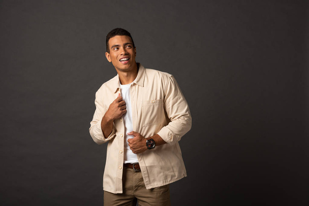 sonriente guapo mixto raza hombre en beige camisa mirando lejos en negro fondo
 - Foto, Imagen