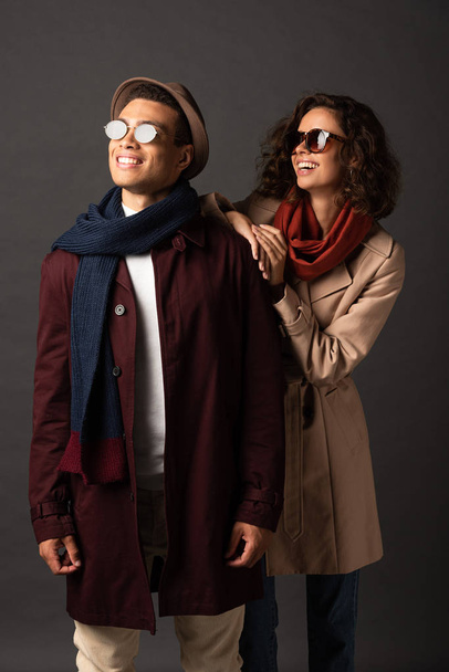 smiling stylish interracial couple in autumn outfit looking away on black background - Foto, afbeelding