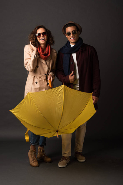 happy stylish interracial couple in autumn outfit with yellow umbrella on black background - Fotoğraf, Görsel