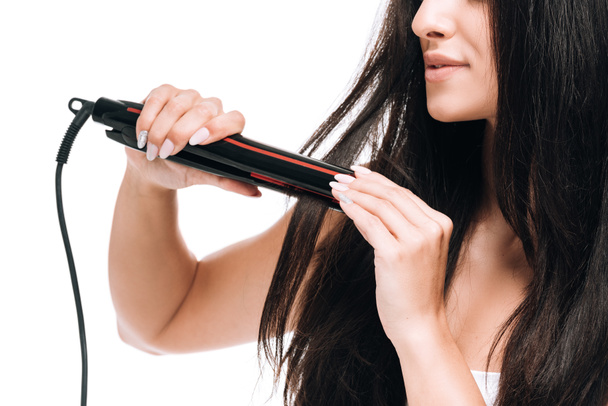 обрезанный вид улыбающейся брюнетки красивая женщина выпрямляя длинные блестящие волосы с плоским железом изолированы на белом
 - Фото, изображение