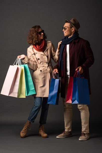 smiling stylish interracial couple in autumn outfit holding shopping bags and looking at each other on black background - Φωτογραφία, εικόνα