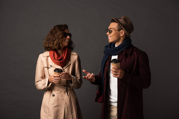 lächelnd stilvolles gemischtrassiges Paar im Herbst-Outfit mit Kaffee zum Reden auf schwarzem Hintergrund - Foto, Bild