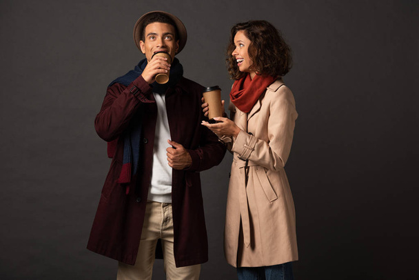 lächelnd stilvolles gemischtrassiges Paar im Herbst-Outfit mit Coffee to go und Gesprächen auf schwarzem Hintergrund - Foto, Bild