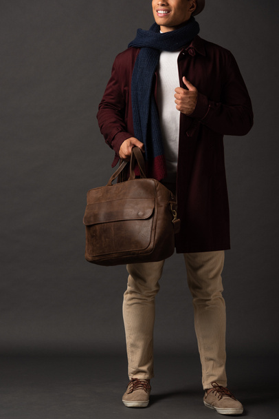 cropped view of smiling mixed race man in scarf and hat holding leather brown bag on black background - Foto, Imagem
