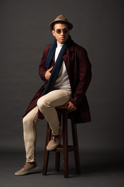 elegant mixed race man in scarf, sunglasses and hat sitting on chair on black background - Φωτογραφία, εικόνα