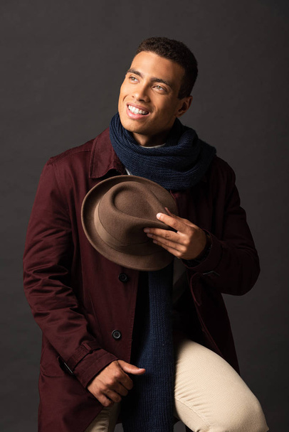 smiling mixed race man in scarf holding hat and looking away on black background - Photo, image