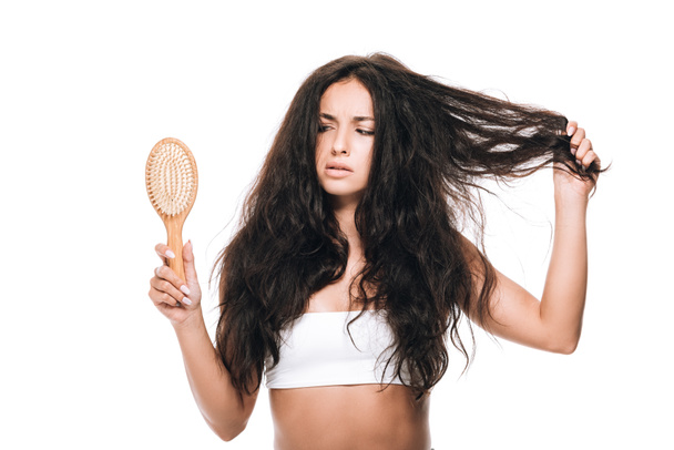 беспокойная брюнетка красивая женщина с волнистыми непослушными волосами глядя на расческу, изолированную на белом
 - Фото, изображение