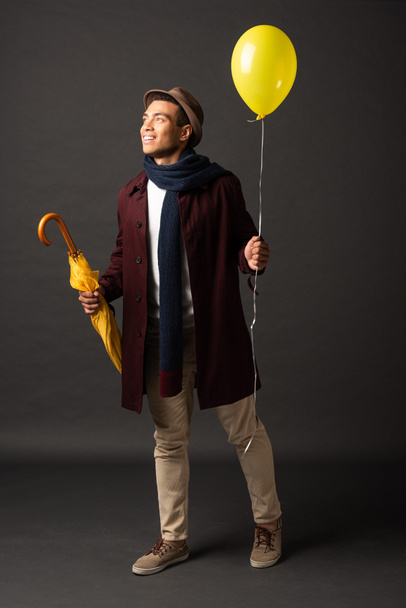 sonriente hombre de raza mixta en bufanda y sombrero sosteniendo globo amarillo y paraguas y mirando hacia otro lado sobre fondo negro
 - Foto, Imagen