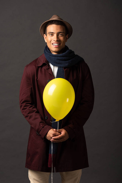 smiling mixed race man in scarf and hat holding yellow balloon on black background - Foto, Imagen