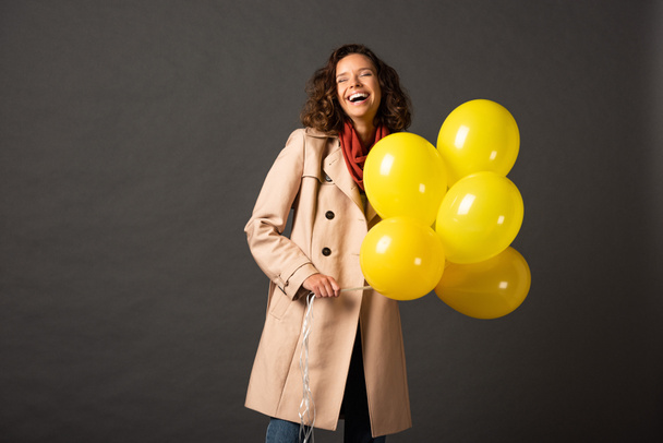 laughing curly woman in trench coat holding yellow balloons on black background - Фото, зображення