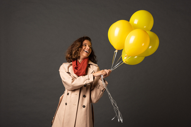 happy curly woman in trench coat holding yellow balloons on black background - Фото, зображення