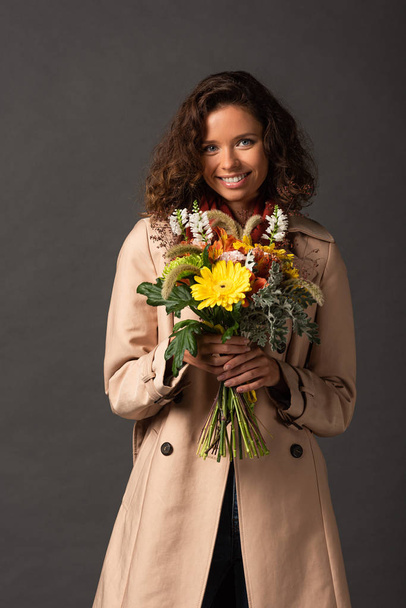 smiling curly woman in trench coat holding bouquet of autumnal wildflowers on black background - Φωτογραφία, εικόνα