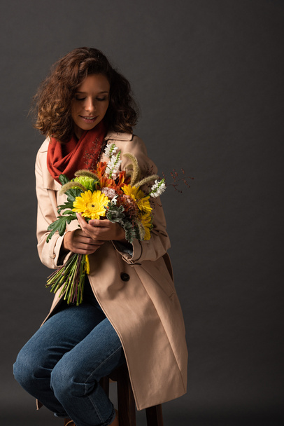 curly woman in trench coat holding bouquet of autumnal wildflowers on black background - Valokuva, kuva