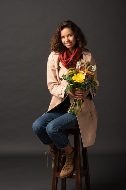 smiling curly woman in trench coat sitting on chair with bouquet of autumnal wildflowers on black background - Foto, Imagen