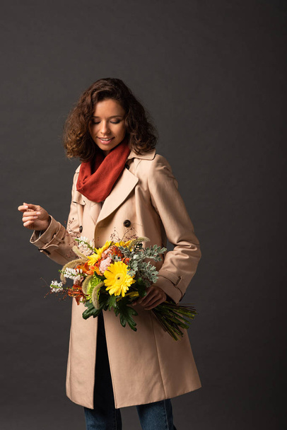 smiling woman in trench coat holding autumnal bouquet on black background - Fotografie, Obrázek