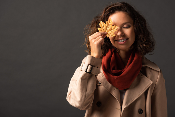 smiling woman in trench coat holding golden maple leaf on eye on black background - Foto, immagini