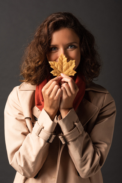 woman in trench coat holding golden maple leaf in mouth on black background - Zdjęcie, obraz