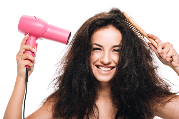усміхнена брюнетка красива жінка укладає кучеряве волосся з рожевим феном і гребінцем ізольована на білому
 - Фото, зображення