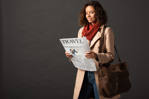 uśmiechnięta kobieta w wykopu płaszcz ze skórzaną torbą trzymając gazety podróży na czarnym tle - Zdjęcie, obraz