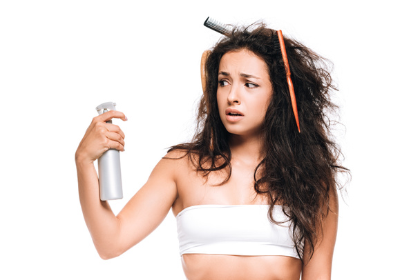 gestresste brünette Frau mit Kämmen im welligen widerspenstigen Haar beim Anblick von Spray isoliert auf Weiß - Foto, Bild