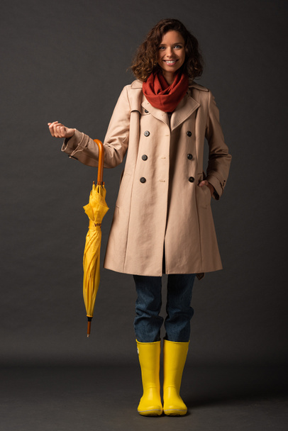 smiling woman in trench coat and rubber boots holding yellow umbrella on black background - Foto, afbeelding