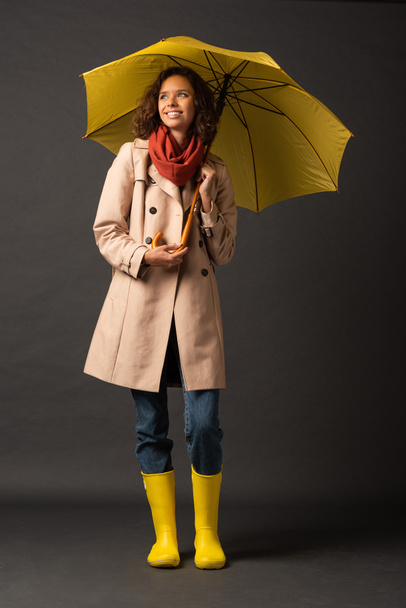 mujer sonriente en gabardina y botas de goma sosteniendo paraguas amarillo y mirando hacia otro lado sobre fondo negro
 - Foto, Imagen