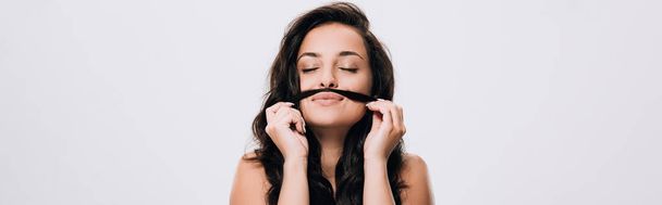 πανοραμικό σφηνάκι μελαχρινή όμορφη γυναίκα με κλειστά μάτια κάνοντας ψεύτικο μουστάκι από μακρά υγιή μαλλιά που απομονώνονται σε γκρι - Φωτογραφία, εικόνα