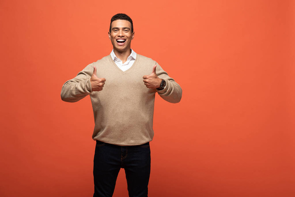 happy mixed race man in beige sweater showing thumbs up isolated on orange - Photo, Image