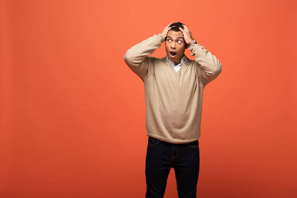 шокированный смешанная раса человек в бежевом свитере держа голову изолированы на оранжевый
 - Фото, изображение