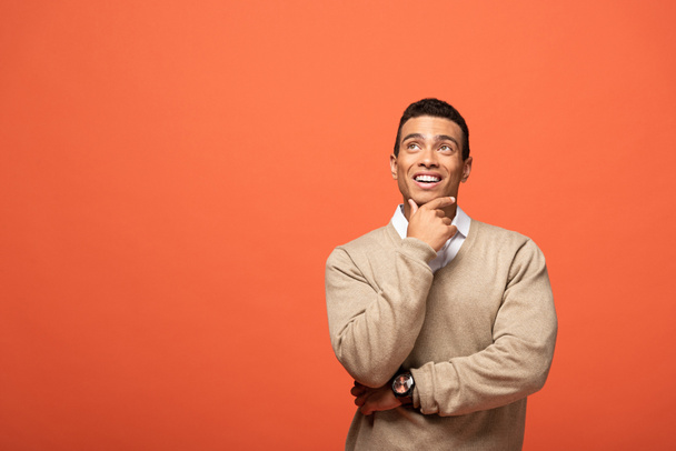 dreamy mixed race man in beige sweater looking away isolated on orange - Фото, изображение