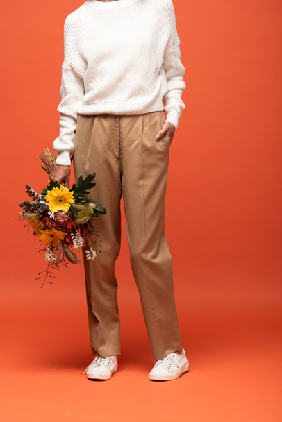 partial view of woman in autumnal outfit with hand in pocket holding bouquet of flowers on orange - Photo, Image