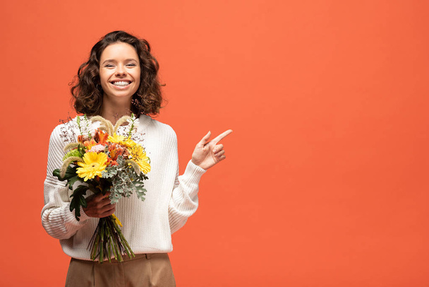 χαρούμενη γυναίκα σε γυναικείο ντύσιμο κρατώντας μπουκέτο λουλουδιών και δείχνοντας με το δάχτυλο απομονωμένο στο πορτοκαλί - Φωτογραφία, εικόνα