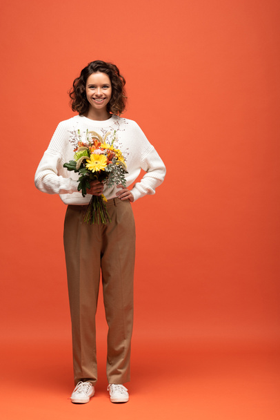 femme heureuse en tenue automnale tenant bouquet de fleurs avec la main sur la hanche sur orange
 - Photo, image