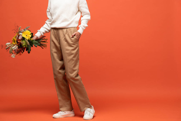 vista recortada de la mujer en traje otoñal con la mano en el bolsillo sosteniendo ramo de flores en naranja
 - Foto, Imagen