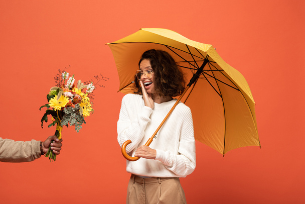 boyfriend gifting shocked woman with umbrella bouquet of flowers isolated on orange - Фото, изображение