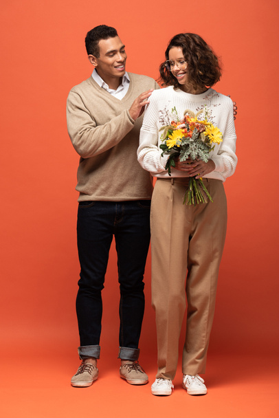 stylish interracial couple in autumn outfit with bouquet of flowers on orange - Foto, Bild