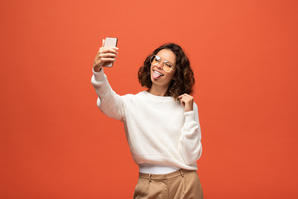 woman in autumnal outfit and glasses showing tongue while taking selfie isolated on orange - Valokuva, kuva