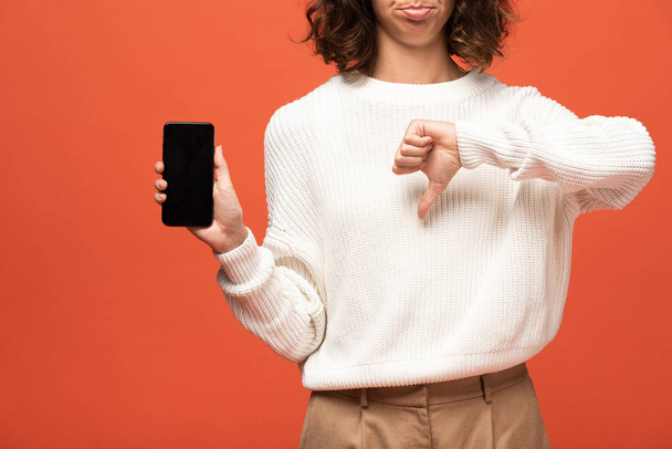 Ausgeschnittene Ansicht einer Frau im herbstlichen Outfit, die ihr Smartphone mit leerem Bildschirm hält und vereinzelt den Daumen nach unten auf orange zeigt - Foto, Bild