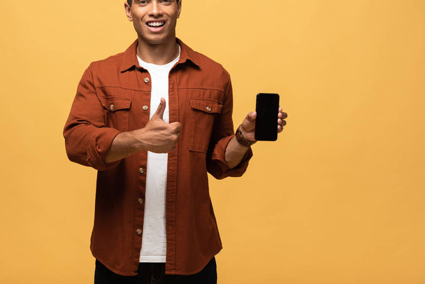 vista recortada de sonriente hombre de raza mixta sosteniendo teléfono inteligente con pantalla en blanco y mostrando el pulgar hacia arriba aislado en amarillo
 - Foto, imagen