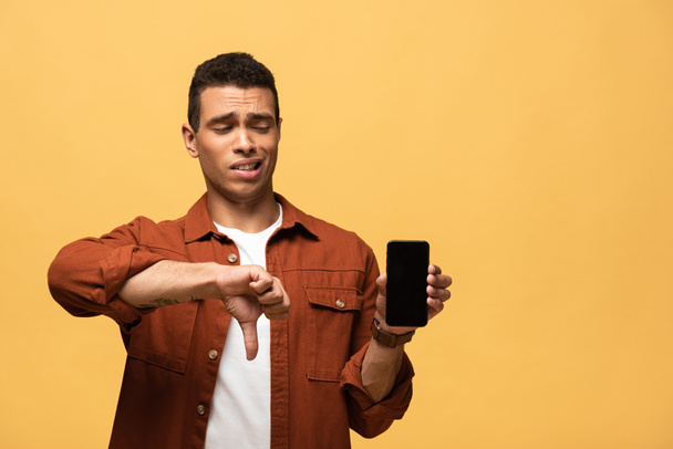 confused mixed race man showing smartphone with blank screen and thumb down isolated on yellow - Фото, зображення