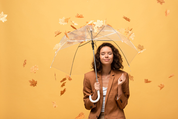 happy curly woman in brown jacket with closed eyes holding umbrella in falling golden maple leaves isolated on yellow - Foto, Imagem