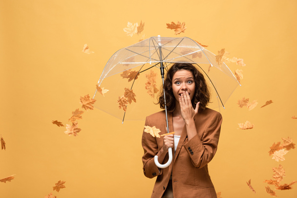 shocked curly woman in brown jacket holding umbrella in falling golden maple leaves isolated on yellow - Photo, Image