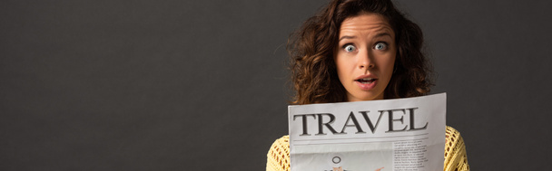 panoramic shot of shocked curly woman in yellow knitted sweater holding travel newspaper on black background - 写真・画像