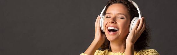 panoramic shot of happy curly woman with closed eyes in yellow knitted sweater listening music in headphones on black background - Photo, Image