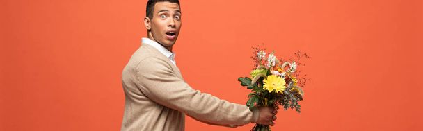 panoramic shot of shocked mixed race man gifting autumnal bouquet isolated on orange - Foto, imagen